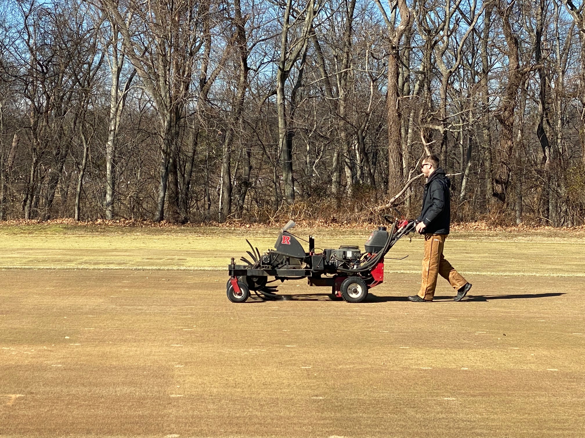 A person walks a wear simulator machine over brown turfgrass research plots.